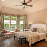 A spacious bedroom with a large bed, two nightstands with lamps, two patterned armchairs by bay windows, a ceiling fan, and wood flooring. Neutral tones and natural light create a cozy atmosphere.