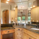 A spacious bathroom with wood cabinets, a green marble countertop, a sink with bronze fixtures, large mirrors, and a walk-in glass shower. Natural light comes through windows with striped valances.