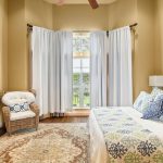 A cozy bedroom with tan walls, a large window with white curtains, a wicker chair with a cushion, a patterned area rug, a blue headboard bed with patterned bedding, and framed art above the bed.