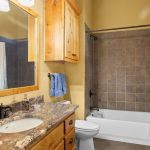 A bathroom with a granite countertop, wooden cabinets, a sink, a toilet, a towel on a rack, and a bathtub with a tiled wall and a white shower curtain. There is a large mirror and two windows with patterned shades.