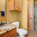 A bathroom with a granite countertop sink, wooden cabinets, a toilet, towel racks with blue towels, and a glass-enclosed shower with beige tile walls and floor.