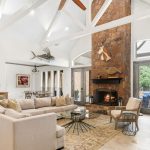Spacious living room with vaulted white ceilings, exposed beams, a large stone fireplace, beige sectional sofa, glass coffee table, and double doors opening to a patio. Animal mounts and modern art decorate the walls.