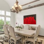 A bright dining room with a rectangular table, six upholstered chairs, a chandelier, large windows, a patterned rug, and a red painting of cats on the white wall. A potted orchid sits on the table.