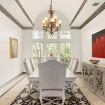 A bright dining room with a long table, eight upholstered chairs, a large window, chandelier, sideboard, modern red artwork, and decorative rug. Light walls and trims contrast with dark door and patterned floor.