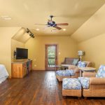 A cozy living space with tan walls and wooden floors features wicker seating with blue patterned cushions, a small bed, a TV on a wooden stand, a ceiling fan, and a door leading to a balcony with a view.