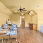 A spacious yellow room with wood floors, wicker armchairs with blue cushions, a bed, wooden cabinets, a ceiling fan, and natural light from a window. Artwork and decorative items are on the tables and cabinets.