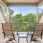 A small balcony with two patio chairs and a table overlooks green trees under a partly cloudy sky, surrounded by beige wooden walls and railing.