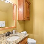 A bathroom with yellow walls, a granite countertop vanity with a sink, a wooden cabinet above the toilet, a large mirror, and two white folded towels hanging on the wall.