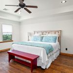 Bright bedroom with a wooden floor, large windows, a bed with white bedding and blue-green pillows, a red bench at the foot, a ceiling fan, a wicker chair, and a wooden shelving unit on the right.