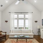 Bright living room with vaulted ceiling, white walls, a sofa and glass coffee table facing built-in shelves and a TV. Two armchairs and a side table sit on a patterned rug near large windows with sheer curtains.