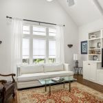 A bright living room with white walls, a white sofa, a glass coffee table, an ornate armchair, built-in shelves with decor and a TV, a large window with sheer curtains, and a red patterned rug on a wooden floor.