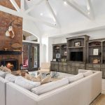 Spacious living room with white sectional sofa, glass coffee table, stone fireplace, mounted deer head, built-in shelves with decor, and large windows overlooking a pool. White vaulted ceiling with exposed beams.