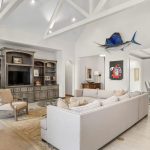 Spacious living room with a large beige sectional sofa, built-in bookshelf and TV, armchair, vaulted ceiling with exposed beams, decorative marlin mounted above a hallway, and a view into a bright dining area.
