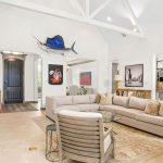 Spacious living room with a large beige sectional sofa, neutral armchairs, wall art, fish mounts, vaulted ceiling with white beams, light tile floors, and an open view into the kitchen and entryway.