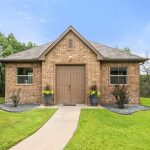 A small brick building with a gable roof, double brown doors, and two windows. Two potted plants sit by the entrance, and a curved walkway leads to the door, surrounded by green lawn and trees.