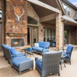 Spacious outdoor patio with wicker furniture and blue cushions, a stone fireplace with a deer head mount, and large windows on a brick house. A garden hose and small plants are visible on the right.
