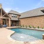 A brick house with a stone-trimmed patio and a swimming pool in the backyard, surrounded by greenery and outdoor seating under a covered area. The sky is partly cloudy.
