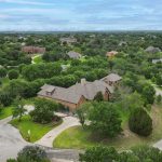 Aerial view of a large house with a stone exterior surrounded by trees and greenery, set in a spacious suburban neighborhood with winding roads and scattered homes.