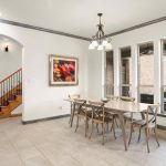 A bright dining area with a wooden table and six chairs, tile flooring, three tall windows, a colorful abstract painting on the wall, and a staircase with wooden steps and railing in the background.