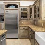 Rustic kitchen with light wood cabinets, granite countertops, stainless steel refrigerator, dishwasher, farmhouse sink, black faucet, and tile backsplash. Large window lets in natural light above the sink.