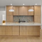 Modern kitchen with light wood cabinets, a large marble island with a built-in sink, stainless steel appliances, and two pendant lights. A dining area with a table and chairs is visible on the right.