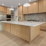 Modern kitchen with light wood cabinets, a large center island with a marble countertop, built-in stainless steel appliances, pendant lights, and light wood flooring. Stairs are visible on the left side.