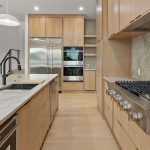 Modern kitchen with light wood cabinets, stainless steel appliances including a double oven and refrigerator, marble countertops, a built-in gas stove, and a large island with a sink and black faucet.