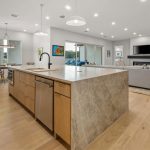 Modern open-concept kitchen and living room with light wood floors, large marble island, pendant lights, neutral-colored furniture, wall-mounted TV, and large windows letting in natural light.