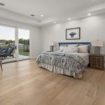 A spacious bedroom with light wood floors, a large bed with patterned bedding, two nightstands with lamps, a lounge chair, and sliding glass doors opening to a patio with lounge chairs and a grassy yard.