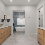 Modern bathroom with double vanities featuring wood cabinets and large mirrors, white countertops, gold fixtures, and a central freestanding tub set against blue marble walls, with sconces and recessed lighting.