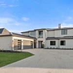Modern two-story house with light-colored exterior, large windows, and a three-car garage. The house has a wide driveway, manicured lawn, and is set against a blue sky with wispy clouds.