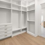 Spacious walk-in closet with white built-in shelves, drawers, and hanging rods, featuring light wood flooring. An open doorway reveals a modern bathroom with a double vanity and mirrors.