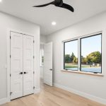 A bright, empty room with light wood floors, white walls, double white doors, a black ceiling fan, and a window overlooking trees and a body of water.