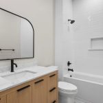 Modern bathroom with a wooden vanity, white countertop, black fixtures, large mirror, white toilet, and a white tiled bathtub-shower combo with built-in shelf.