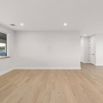 A bright, empty room with light wood floors, white walls, recessed ceiling lights, a large window overlooking a river, and French doors leading to another area.