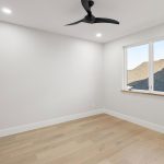 A bright, empty room with light wood floors, white walls, a modern black ceiling fan, recessed lighting, and a window showing rooftops outside.