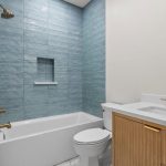 Modern bathroom with a bathtub and shower, blue tiled wall, built-in shelf, white toilet, white countertop, wood vanity, gold fixtures, and white walls.
