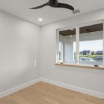 A small, empty room with light wood flooring, white walls, a black ceiling fan, recessed ceiling lights, and a large window overlooking houses and a body of water.