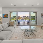 A modern living room with a light gray sofa, glass coffee table, and rug, opening onto a patio with a pool and outdoor dining area. Large sliding glass doors provide a view of houses and greenery outside.