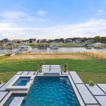 A backyard with a rectangular swimming pool and hot tub, next to a lawn and fenced area, overlooks a calm lake with docks and waterfront houses under a partly cloudy sky. Two blue lounge chairs are by the pool.