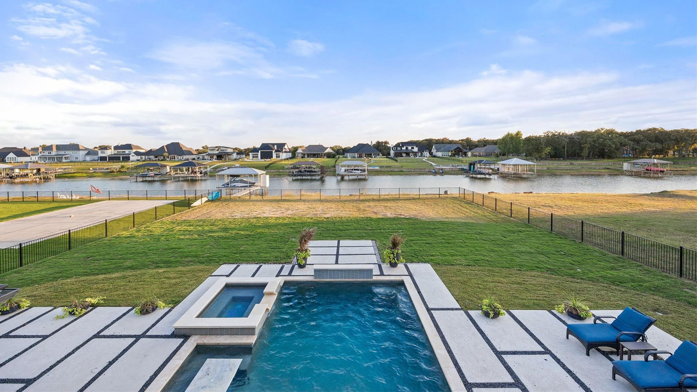 A backyard with a rectangular swimming pool and hot tub, next to a lawn and fenced area, overlooks a calm lake with docks and waterfront houses under a partly cloudy sky. Two blue lounge chairs are by the pool.