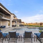Modern backyard with a swimming pool, lounge chairs, and a spacious patio area next to a two-story house. Green lawn and neighboring houses are visible in the background under a blue sky.