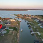 Aerial view of a lakeside neighborhood with houses along canals leading to a large lake. A red location marker highlights one house near the water’s edge. Lush green lawns and docks are visible.