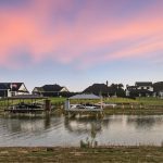 A serene lakeside scene at sunset with modern houses, private docks, and boats along the shore; pastel pink and purple clouds fill the sky, reflecting peacefully on the water.