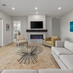 Modern living room with light walls and a large sectional sofa, glass coffee table, fireplace under a wall-mounted TV, beige armchair, and a colorful fish painting. Sliding glass doors open to a patio area.