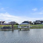 Waterfront homes with boat docks and covered patios line a grassy shoreline under a blue sky with scattered clouds. The calm water reflects the houses and docks.
