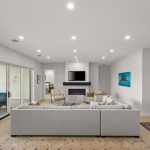 A modern living room with a large gray sectional sofa, a fireplace with a TV above it, wall art, a rug, and sliding glass doors leading to a patio with outdoor seating and a pool.