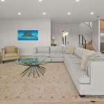 A modern living room with a large gray sectional sofa, glass coffee table, beige armchair, wall-mounted TV, electric fireplace, and open kitchen with light wood cabinets and stainless steel appliances. A fish artwork hangs on the wall.