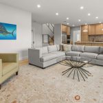 Modern living room with a large light gray sectional sofa, a green armchair, a glass coffee table, and a patterned rug. An abstract fish painting hangs on the wall, and an open kitchen is visible in the background.