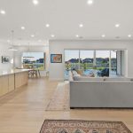 Bright open-plan living area with a modern kitchen, large island, light wood cabinets, dining table, gray sectional sofa, fireplace, and floor-to-ceiling windows overlooking a patio and waterfront view.